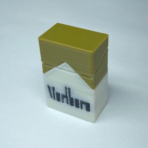 May include: A small, rectangular, 3D-printed object shaped like a cigarette pack. The top section is a solid mustard yellow, while the bottom section is white with the black text 'Marlboro' printed across the front in a stylized, blocky font.