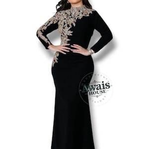 Pode incluir: Um vestido preto até ao chão com mangas compridas e decote alto. O corpete e a manga direita apresentam um intrincado bordado floral adornado com lantejoulas prateadas e douradas. Um logótipo circular com o texto Awais House é visível.