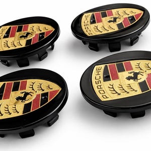 May include: Four black wheel center caps with the Porsche logo. Each cap features a gold emblem with the Porsche name, a black horse, and red and black stripes. The caps are oval-shaped and have a glossy finish.