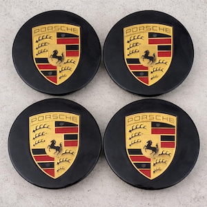 May include: Four black Porsche wheel center caps. Each cap features the Porsche crest in gold, red, and black, with the word "PORSCHE" above. The crest includes a black horse and a shield design. The caps are circular.