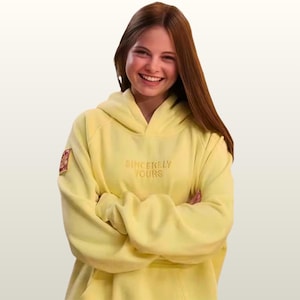 May include: A pale yellow hoodie featuring the embroidered words "Sincerely Yours". The hoodie has a hood and a small patch on the left sleeve. The person in the image is smiling with their arms crossed.
