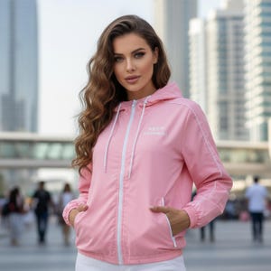 May include: A light pink hooded jacket with white trim and a white zipper. The jacket has the words "KEEP SWIMMING" printed on the chest. The jacket has a drawstring hood and pockets. The model is wearing white pants.