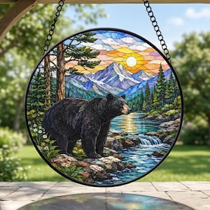 Peut inclure: Un capteur de soleil circulaire en vitrail représentant un ours noir debout sur une rive rocheuse. La scène comprend un ruisseau, une cascade, des pins et des montagnes enneigées sous un ciel au coucher du soleil avec un soleil jaune.