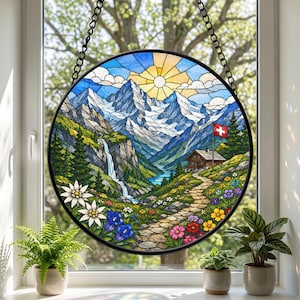 May include: A circular stained glass suncatcher featuring a mountain landscape with snow-capped peaks, a waterfall, a stone path, and a wooden cabin with a Swiss flag. The scene includes colorful wildflowers, green pine trees, and a yellow sun.