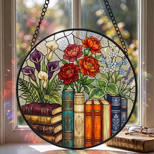 May include: A round stained glass window featuring a floral design with red, orange, and purple flowers, and a stack of books. The window is framed in black and hangs from a chain. The flowers are in vases that look like books.