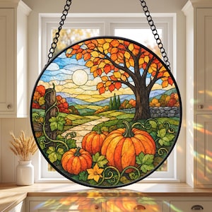 May include: A circular stained glass suncatcher featuring an autumn landscape with orange pumpkins in the foreground, a winding stone path, a large tree with golden leaves, and a pale sun in a blue sky, all framed in a black metal border with a chain.