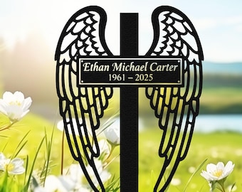 May include: Black metal memorial cross with angel wings. The cross features the name "Ethan Michael Carter" and the years "1961-2025". The wings are detailed with feather patterns. The background shows a field of white flowers and a blurred landscape.