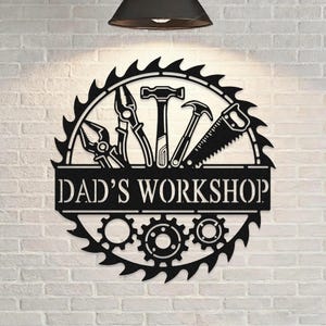 May include: Black metal sign with the text "DAD'S WORKSHOP" and a circular saw blade design. The sign features silhouettes of tools including pliers, a hammer, a saw, and gears, perfect for a workshop or garage.