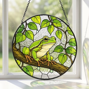 May include: A circular stained glass suncatcher featuring a bright green frog perched on a brown tree branch. The frog is surrounded by vibrant green leaves and a textured clear glass background. The piece is framed in black and hangs by a chain.