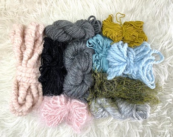 May include: Assortment of yarn in various colors and textures, including pink, black, gray, blue, and green. The yarn is arranged on a white fluffy surface.