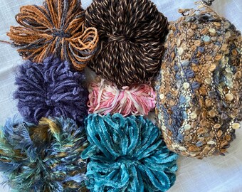 May include: A collection of yarn in various colors and textures, including brown, blue, teal, pink, and gray. The yarn is arranged in a circle on a white surface.