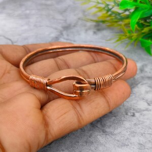 Op de afbeelding: Een koperkleurige metalen armband met een haaksluiting. De cirkelvormige band is gemaakt van een enkel stuk draad dat terugloopt om de sluiting te vormen, met aan beide zijden gedetailleerde wikkelingen van metaaldraad.