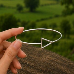 May include: A silver-toned metal bangle bracelet featuring a thin, circular band that terminates in an open triangular shape. The bracelet is constructed from a smooth, polished wire material and maintains a consistent, simple geometric design.