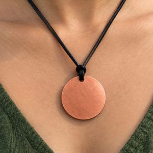 May include: A circular copper pendant with a smooth, matte finish suspended from a simple black leather cord. The pendant features a single hole at the top where the cord is threaded through, creating a clean, minimalist look against the skin.