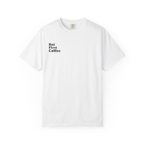 May include: A white short-sleeved t-shirt with a crew neckline. The front left chest area features the black text "But First Coffee" printed in a bold, rounded serif font. The garment has a standard fit with visible stitching at the hem and sleeves.
