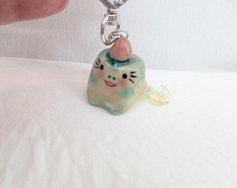 May include: A whimsical keychain featuring a small, square-shaped character with a smiling face, black whiskers, and a pink strawberry on top. The character is light blue and cream-colored, attached to a silver heart-shaped clasp.