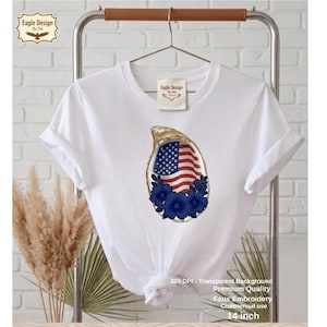 Puede incluir: Camiseta blanca con un diseño que presenta una concha de ostra con una bandera estadounidense y flores azules. La camiseta mide 35,56 cm de largo. El texto de la camiseta dice "Eagle Design by Ella".