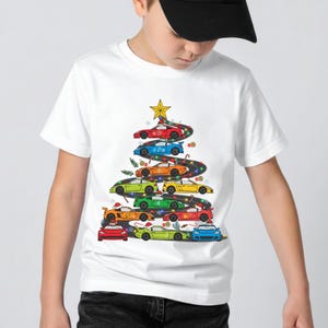 Könnte beinhalten: Ein weißes Kurzarm-T-Shirt mit einer Grafik eines Weihnachtsbaums, der aus einer kurvenreichen Straße besteht, auf der verschiedene bunte Sportwagen geparkt sind. Der Baum ist mit einem gelben Stern sowie bunten Lichterketten und Süßigkeiten verziert.