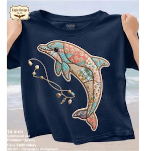Peut inclure: T-shirt bleu marine avec un motif de dauphin coloré et un motif patchwork. Le dauphin porte un collier de perles et est orné d'éléments sur le thème de la mer. Le t-shirt mesure 35,5 cm.