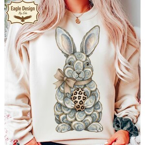 May include: Cream-colored sweatshirt featuring a watercolor illustration of a bunny holding an Easter egg. The bunny is gray and white with a tan bow. The egg has a leopard print design. The sweatshirt has the text "Eagle Design By Eda" in a decorative logo.