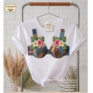 Op de afbeelding: Wit T-shirt met een faux geborduurd bh-ontwerp. De bh heeft denim, geruite en bloemenaccenten met roze en gele bloemen. Het shirt hangt aan een metalen rek. De tekst op het shirt luidt "Eagle Design by Ella".