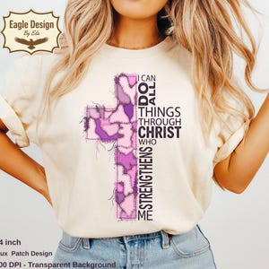 May include: A cream-coloured t-shirt with a cross design made of pink and purple patches. The cross has the text "I CAN DO ALL THINGS THROUGH CHRIST WHO STRENGTHENS ME". The shirt is 35.56 cm in length.