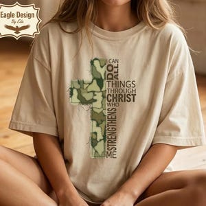 Op de afbeelding: Beige T-shirt met een kruisontwerp van camouflage stof. Het kruis staat centraal op het shirt en de tekst "I CAN DO ALL THINGS THROUGH CHRIST WHO STRENGTHENS ME" is verticaal aan de rechterkant gedrukt.