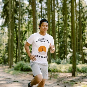 May include: A person running on a path in a forest, wearing a white t-shirt with the text "CAMINO DE SANTIAGO -2026" and a graphic of a cross and sun. They are also wearing gray shorts, a black cap, and black sneakers.