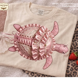 以下が含まれることがあります： クリーム色のTシャツ。ピンク色のカメのデザインで、コルセット風の甲羅、レースのディテール、フリル付きトリムが特徴です。「Eagle Design by Eda」と「COMFORT COLORS」の文字が含まれています。「300 DPI - Transparent Background Commercial use 35.56 cm」の文字も含まれています。