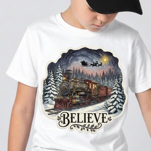 Peut inclure: T-shirt blanc avec une illustration colorée d'un train vintage traversant une forêt enneigée sous un ciel étoilé. L'image comprend le mot "BELIEVE" dans une police décorative.