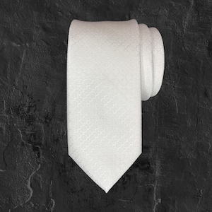 Op de afbeelding: Een witte stropdas met een subtiel, herhalend geometrisch patroon. De stropdas is netjes opgerold en gepresenteerd tegen een donkere, getextureerde achtergrond. De stof van de stropdas heeft een lichte glans.