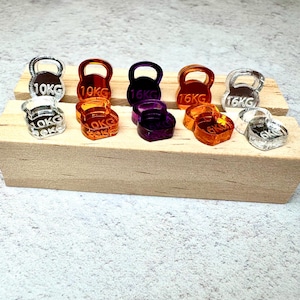 May include: A selection of miniature kettlebell charms in shades of clear, orange, purple, and silver. Each charm is marked with its weight, either 10KG or 16KG, and is presented on a small wooden platform.