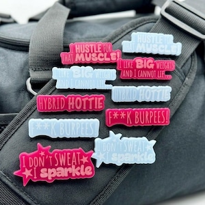May include: A collection of colorful, rectangular gym bag charms with motivational phrases like "Hustle for that muscle" and "I don't sweat, I sparkle." The charms are in shades of pink and blue, and are placed on a black gym bag.
