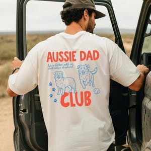 May include: Cream-colored t-shirt with the text "AUSSIE DAD" in red, and "CLUB" in red. Below the text are two blue line-drawn Australian Shepherds, paw prints, and a tennis ball. The shirt also has the text "life is better with an Australian shepherd."