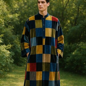 Puede incluir: Una larga bata de terciopelo multicolor con un patrón de tablero de ajedrez. La bata presenta un cuello mandarín, mangas largas y bolsillos. La paleta de colores incluye tonos de azul, amarillo, rojo y negro. La persona está de pie al aire libre en un entorno natural verde.