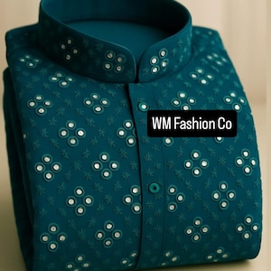 Puede incluir: Camisa de color turquesa con cuello mandarín y bordados detallados. La camisa tiene una botonadura frontal y pequeños adornos reflectantes. El texto "WM Fashion Co" se muestra en la parte delantera.