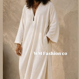 May include: A long-sleeved, floor-length off-white robe featuring a V-neckline, side pockets, and high side slits. The garment displays the text WM Fashion co on the front. The fabric has a textured appearance, and the design includes a simple cut.