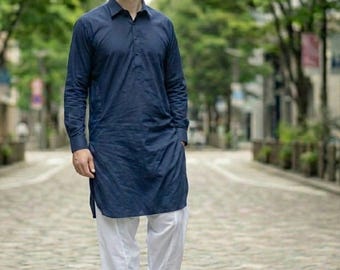 May include: A navy blue long-sleeved tunic shirt with a mandarin collar, paired with white, loose-fitting trousers. The outfit is completed with brown leather sandals. The person is walking on a cobblestone path.