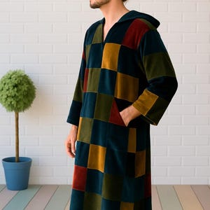 Puede incluir: Una bata larga con capucha con un dise&ntilde;o de patchwork en tonos de verde azulado, amarillo mostaza, rojo y verde oliva. La bata tiene mangas largas y un bolsillo delantero. La persona est&aacute; de pie frente a una pared de ladrillos blancos.