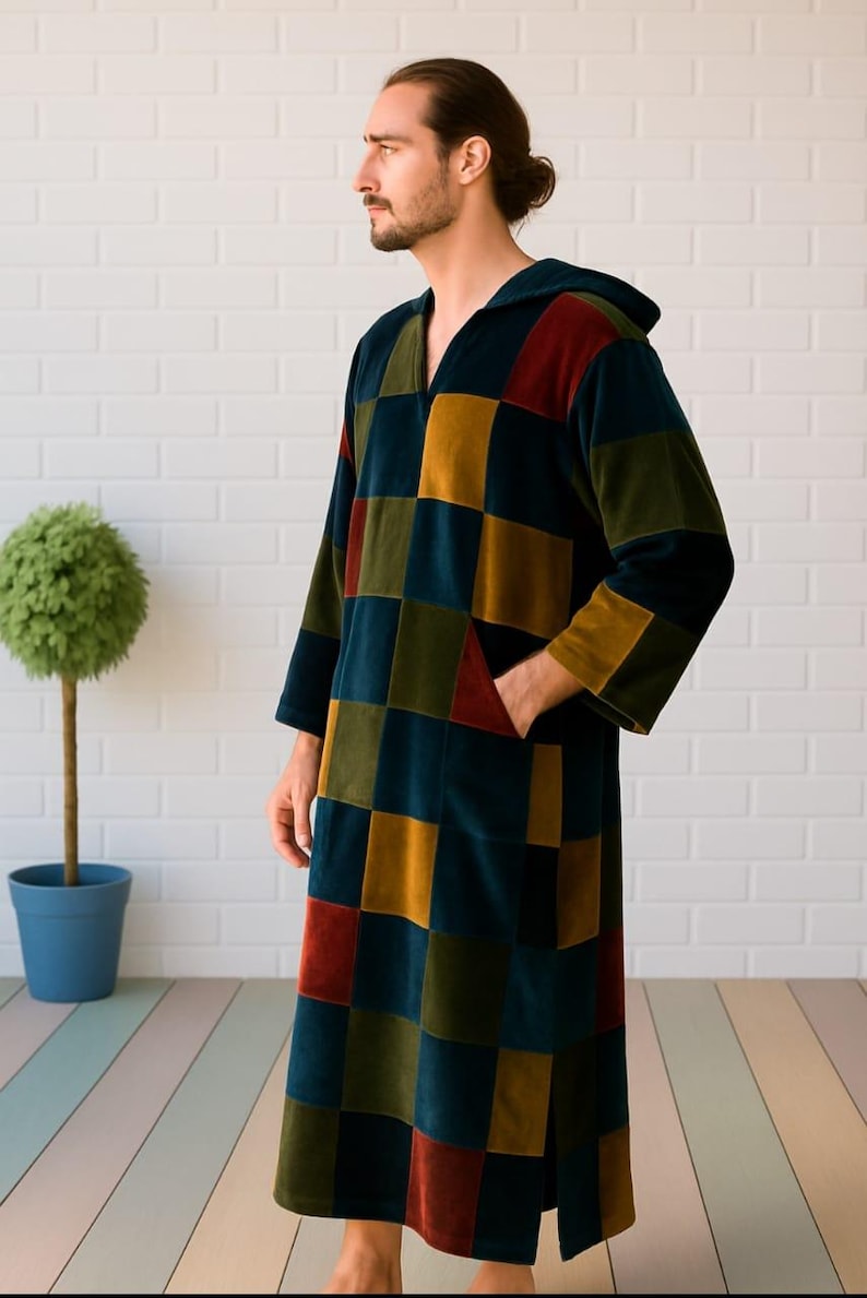 Puede incluir: Una bata larga con capucha con un dise&ntilde;o de patchwork en tonos de verde azulado, amarillo mostaza, rojo y verde oliva. La bata tiene mangas largas y un bolsillo delantero. La persona est&aacute; de pie frente a una pared de ladrillos blancos.