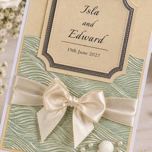 May include: A rectangular wedding invitation card featuring a cream-coloured top section with the text Isla and Edward 19th June 2027, a sage green textured wave-patterned bottom section, a cream satin ribbon bow, and small white shell and pearl accents.