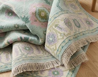 Puede incluir: Una alfombra rectangular plegada con un estampado floral en tonos de azul claro, rosa y morado. La alfombra tiene un borde crema y flecos en los bordes. Está sobre un suelo de madera.