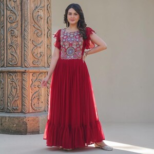 May include: A floor-length deep red dress featuring short ruffled sleeves and a round neckline. The bodice is adorned with a dense, multi-colored circular embroidery pattern in shades of blue, gold, and white. The skirt is pleated and flows.