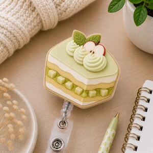 May include: A badge reel featuring a three-dimensional silicone charm shaped like a slice of layered cake. The cake is light green and cream-colored, topped with piped frosting, two green leaves, and a small red apple slice. A clear plastic strap is attached.