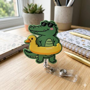 May include: A badge reel featuring a green cartoon alligator wearing black sunglasses and holding a yellow duck-shaped inflatable swim ring. The alligator has a light green belly and a smiling expression. The reel has a clear plastic strap and clip.