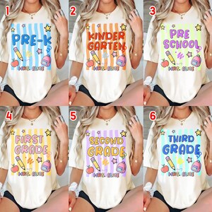 以下が含まれることがあります： クリーム色のTシャツで、カラフルな学校をテーマにしたデザインです。各シャツには、Pre-K、幼稚園、プレスクール、1年生、2年生、3年生の学年が表示されています。デザインには、鉛筆、リンゴ、バックパック、星が含まれています。「MRS. ALICE」の文字も含まれています。