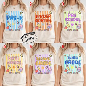 以下が含まれることがあります： 学校をテーマにしたグラフィックが特徴のアイボリー色のTシャツ。各シャツは、Pre-K、幼稚園、プレスクール、1年生、2年生、3年生など、異なる学年を表しています。デザインには、鉛筆、リンゴ、バックパック、星が含まれています。