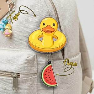 May include: A yellow duck-shaped pool float topper and a watermelon slice charm are attached to a backpack. The duck float has a round shape with a smiling face. The watermelon charm is red with black seeds.