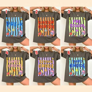 以下が含まれることがあります： 学校をテーマにしたカラフルなデザインの濃い灰色のTシャツ6枚。各シャツには、Pre-K、幼稚園、プレスクール、1年生、2年生、3年生の学年が表示されています。デザインには、鉛筆、リンゴ、バックパックが含まれています。