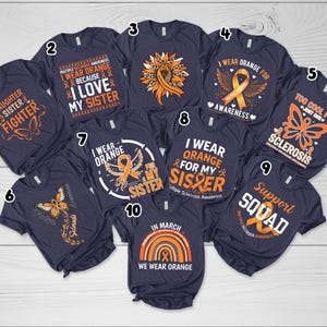 May include: A collection of navy blue t-shirts with various orange and white designs. The shirts feature text and graphics related to Multiple Sclerosis awareness, including butterflies, ribbons, and phrases like "I wear orange."